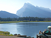 Woman relaxes on front of truck by lake and mountains