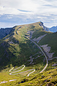 Steile Straße der Kurven auf grünen Wiesen umrahmt von felsigen Gipfeln Sorland Vaeroy Island Grafschaft Nordland Lofoten Norwegen Europa