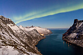 Northern Lights (aurora borealis) auf den schneebedeckten Gipfeln und dem Dorf Fjordgard umrahmt von dem gefrorenen Meer, Ornfjorden, Senja, Troms, Norwegen, Skandinavien, Europa