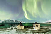 The Northern Lights (aurora borealis) and stars illuminate the wooden huts by icy sea, Djupvik, Lyngen Alps, Troms, Norway, Scandinavia, Europe