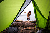 A shirtless man swings a large stick like a baseball bat on a remote beach, as seen through the opening in a green tent door.
