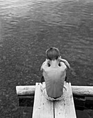 Rear View Portrait of Sad Boy Sitting Hugging His Knees at End of Dock on Lake, High Angle View