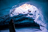 A man looks out an opening in the ceiling of an ice cave within Augustana Glacier in the winter, Alaska Range, Interior Alaska, USA