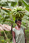 Ein Porträt einer Frau lächelt und trägt eine große Gruppe von Bananen auf dem Kopf, Uganda, Afrika