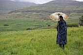 A man holding an umbrella and wrapped in a blue blanket stands in the mountains, Lesotho, Africa