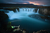 Human figure with hood and headlamp on sitting on the edge of huge waterfall during the night. Low clouds reflect light from volcanic activity in the distance.