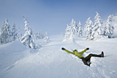 Man lying happily in deep snow on an alpine plateau surrounded by snow covered spruce trees, looking towards the sky. He's dressed with a green waterproof jacket, waterproof pants, mittens, gaiters and winter trekking boots.