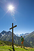 Ein männlicher Wanderer geht zu einem hölzernen Kreuz auf einer Bergwiese im Val d'Anniviers in den Schweizer Alpen. Die Sonne scheint hell, die Temperaturen sind hoch. Dies ist auf halbem Weg die Haute Route, eine klassische Wanderung zwischen Chamonix i