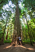 Eine junge Frau umarmt eine alte Douglasie im Cathedral Grove, Cathedral Grove, British Columbia.