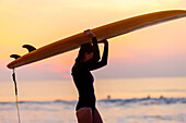 Female surfer carrying board above head at sunrise