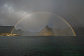 Regenbogen scheint in Reinefjord über Olstind Berggipfel, Toppøy, Lofoten Inseln, Norwegen