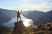 A female hiker in a red shirt celebrates at the top of the Mount Storm King trail in Olympic National Park overlooking Lake Crescent.