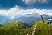 Distant View Of Mountain Bike On Summit Of Mountain Lifting Mountain Bike Above His Head