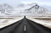 Die schwarze Asphaltstraße, die in Richtung zum Schnee führt, bedeckte Berge im Winter, Südisland, polare Regionen