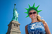 young american tourist wearing a styrofoam crown posing in front of the statue of liberty, new york harbor, new york city, state of new york, united states, usa