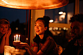 Happy woman holding small birthday cake with lit candles while sitting by friends at home