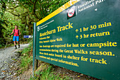 Wanderwegweiser für Routeburn Track, Frau beim Wandern im Hintergrund, Routeburn Track, Great Walks, Fiordlands Nationalpark, UNESCO Welterbe Te Wahipounamu, Queenstown-Lake District, Otago, Südinsel, Neuseeland