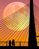 Silhouettes of people at sunset, walking near the Zakim Bridge in Boston, Massachusetts, as the full moon rises