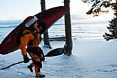 Kayak surfer carrying a paddle and kayak in winter along the snow covered shore, Homer, Southcentral Alaska, USA