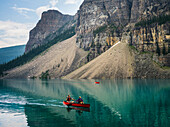 A red canoe in Moraine Lake with a cliff of the Canadian Rocky Mountains along the shoreline; Lake Louise, Alberta, Canada