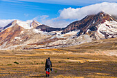 A backpacker crosses the exotic landscape of the ash and pumice-covered Valley of Ten Thousand Smokes in Katmai National Park, with Mt. Katmai (left), Trident Volcano (right), and the Knife Creek Glaciers looming in the distance; Alaska, United States of 