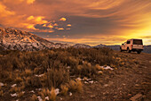 Small camper truck stopped in desert scenery with snow-capped mountains in the background. The sunrise lights the mountains and the clouds with bright red colours; Potrerillos, Mendoza, Argentina