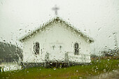 A white church building with cross viewed through a window wet with raindrops; Newfoundland, Canada
