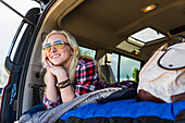A young woman on a road trip lays in the back of a vehicle with a sleeping bag looking out from the  open door; Edmonton, Alberta, Canada