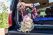 A young woman on a road trip lays in the back of a vehicle with a sleeping bag using her smart phone; Edmonton, Alberta, Canada