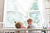 Two sisters, a baby and a toddler, play together in a crib by a window; Sorrento, British Columbia, Canada