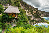 Woman sitting outside thatched roof hut on coast with cliffs, Nusa Penida, Bali, Indonesia