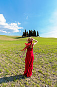 San Quirico d'Orcia, Orcia valley, Siena, Tuscany, Italy, A young woman in red dress admiring the view in a wheat field near the cypresses of Orcia valley