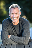 Portrait of gray-haired man smiling and leaning on outdoor table with crossed arms, Massachusetts, USA