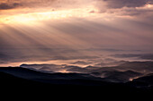 Sun shining through clouds over silhouettes of hills shrouded in morning fog, Linville, North Carolina, USA
