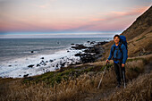 Male backpacker standing with trekking poles at shore of Lost Coast at dusk, California, USA