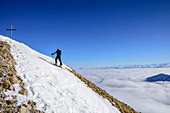 Mann auf Skitour steigt zum Gipfelkreuz des Hinteren Sonnwendjoch auf, Nebelmeer im Tal, Hinteres Sonnwendjoch, Bayerische Alpen, Tirol, Österreich