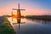 Windmill reflected in the canal framed by grass and pink sky at dawn, North Holland, The Netherlands