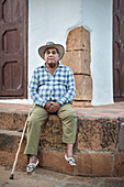 alter Kolumbianischer Mann (Einheimischer) mit Stock und Hut wurde vor einem typischen kolonialen Gebäude sitzend porträtiert, Barichara, Departmento Santander, Kolumbien, Südamerika