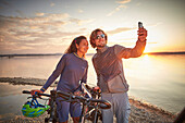 Young  woman on touring bike, young man on touring eBike on tour,taking selfie on  lakeshore, Muensing, bavaria, germany