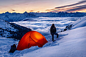 Young woman is standing next to an orange, enlightet tent in the snow, sea of clouds, wintery mountains, Innsbruck, Tyrol, Austria