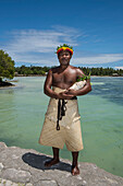 Ein Mann mit Blatt-und-Blumenkrone und Körperbesatz aus Pflanzenmaterial steht in der Nähe des Wassers und hält eine große Muschel, Butaritari Atoll, Gilbert-Inseln, Kiribati, Südpazifik