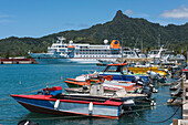 A long row of brightly colored boats are tied to a pier with expedition cruise ship MS Bremen (Hapag-Lloyd Cruises) in the background, Rarotonga, Cook Islands, South Pacific