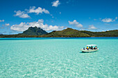 Ein kleines Motorboot mit Touristen fährt über relativ flachem, hellgrünem Wasser in der Lagune, mit der Insel und den hohen Bergen im Hintergrund, Bora Bora, Gesellschaftsinseln, Französisch-Polynesien, Südpazifik