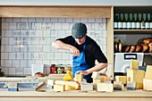 Käsehändler schneidet Käse mit Käsedraht, umgeben von einer Vielzahl von Käsesorten auf der Arbeitsplatte