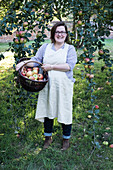 Woman wearing apron holding brown wicker basket with freshly picked apples, smiling at camera.