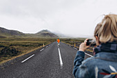 Young woman photographing boyfriend walking on highway in Iceland
