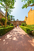 People on garden path leading to the entrance of Real Alcazar. The complex is a mix of Mudejar, Islamic, Gothic, Renaissance and Baroque architectural style. Seville, Andalusia, Spain