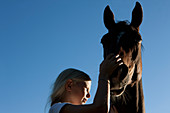 Girl petting muzzle of horse under blue sky
