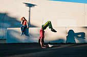 Young woman watching man do handstand along urban wall