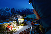 Young woman lies in her bivouac sack on a sleeping pad in the snow and boils water with a gas cooker in the glow of her headlamp in front of the Karwendel backdrop, Tyrol, Austria